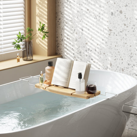 White bathtub with a wooden bath caddy holding an open book and toiletries under a window